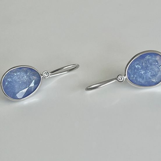 Zwei silberne Ohrringe mit blauen Edelsteinen, jeweils mit einem kleinen Diamanten verziert, ruhen auf einer weißen Oberfläche.