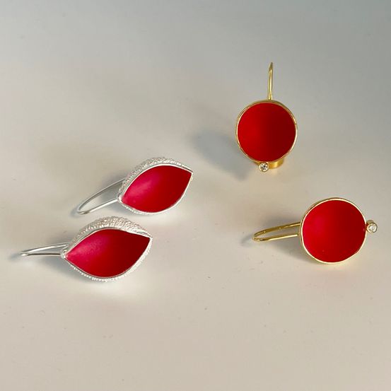 Vier Schmuckstücke, zwei Ohrringe und zwei Ringe, sind ausgestellt. Die Ohrringe haben rote Blütenblätter mit silbernen Details, während die Ringe rote Zentren mit einem einzelnen goldenen Diamanten haben.