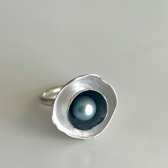 Ein silberner Ring mit einer zentralen, glänzenden blauen Perle, vor einem matten grauen Hintergrund.