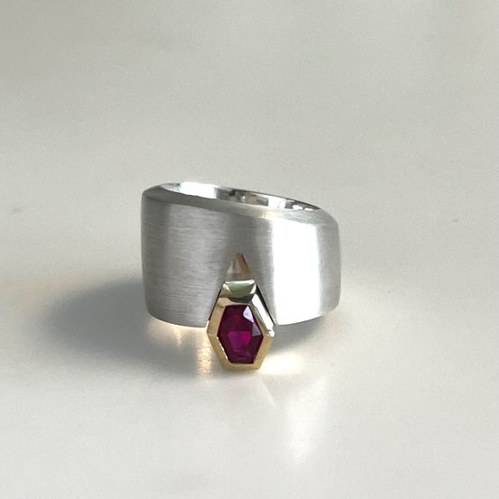 Ein silberner Ring mit geometrischem Design zeigt einen roten Edelstein in der Mitte, montiert auf einer goldenen Basis. Der Ring präsentiert ein modernes Design.