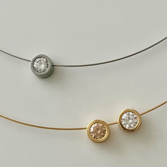 Zwei Halsketten, eine mit einer silbernen Kette und einem einzelnen Diamanten, die andere mit einer goldenen Kette und zwei Diamanten, auf einer weißen Oberfläche ausgestellt.