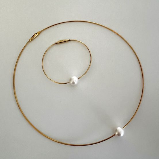 Eine goldene Halskette mit einer weißen Perlenkette ruht auf einer weißen Oberfläche. Die Halskette besteht aus einem größeren, goldenen Ring mit einem kleineren, perlenverzierten Ring darin.