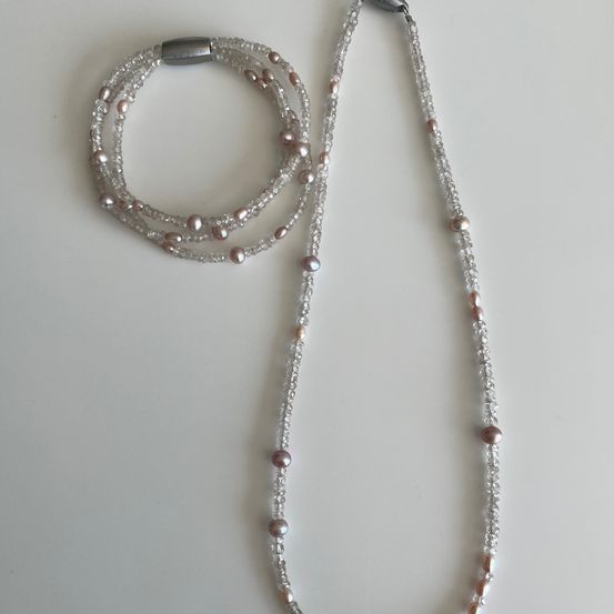 Ein Schmuckset mit einer langen Kette und einem Armband aus rosa und silbernen Perlen, das auf einer weißen Oberfläche präsentiert wird.