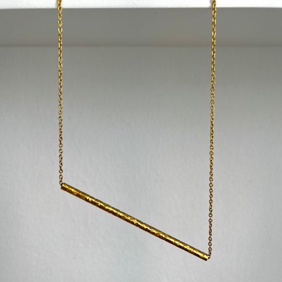 Eine goldene Kette mit einem geraden Stab-Anhänger hängt von einer weißen Oberfläche. Der Stab hat eine leicht gekrümmte Textur.