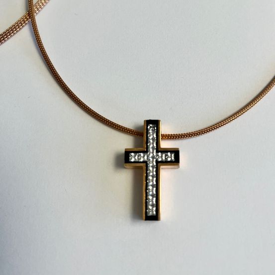 Ein goldener Kreuzanhänger an einer goldenen Kette, auf einer weißen Oberfläche platziert. Das Kreuz ist mit schwarzen und weißen Diamanten verziert.