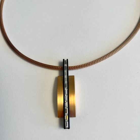 Eine braune Halskette mit einem Anhänger aus Messing und Diamanten. Der Anhänger ist auf einem schwarzen Balken montiert. Die Halskette ist auf einem weißen Hintergrund ausgestellt.