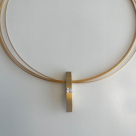 Ein goldener Anhänger mit einem Diamanten liegt auf einem weißen Hintergrund. Der Anhänger hat eine rechteckige Basis und einen kleinen Diamanten in der Mitte eingebettet.