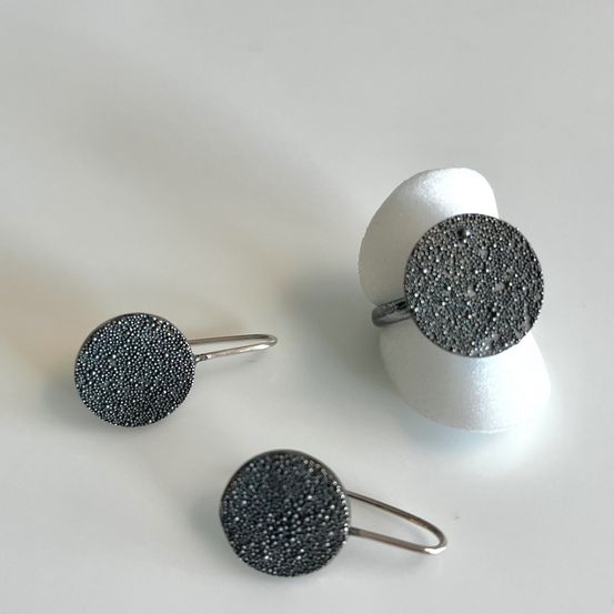 Drei runde Ohrringe mit einem schwarzen und silbernen Punktmuster sind auf einer weißen Oberfläche zu sehen. Zwei sind kleiner und haben Metallschlaufen. Der dritte ist größer und befindet sich auf einem weißen Objekt.