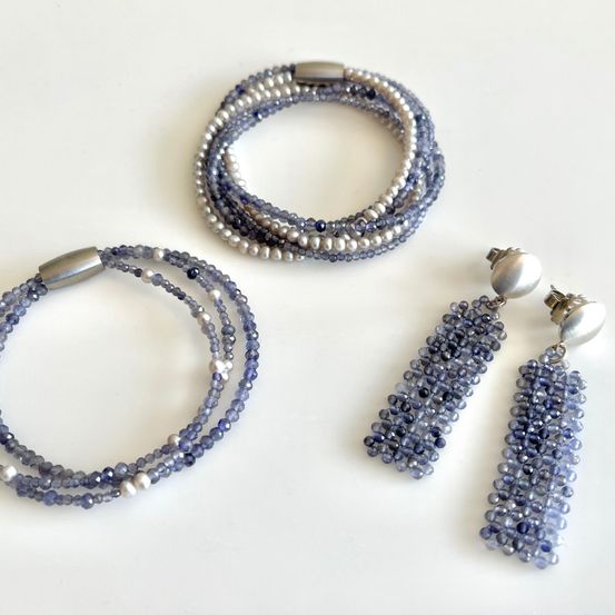 Drei blaue Perlenschmuckstücke sind auf einer weißen Oberfläche ausgestellt. Das erste ist ein Armband, gefolgt von einem Stapel geschichteter Armbänder. Der dritte Gegenstand ist ein Paar hängende Ohrringe.
