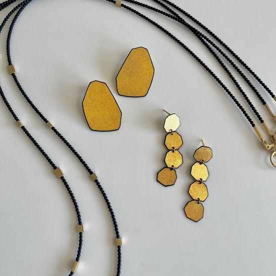 Ein Set aus goldenen hexagonalen Anhängern und passenden Ohrringen mit schwarzperliger Kette auf einer weißen Oberfläche.
