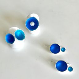 Vier Stücke blau emaillierter Schmuck auf einer weißen Oberfläche. Ein Paar Ohrringe mit einem großen blauen Kreis und einem kleineren blauen Kreis darunter. Ein anderes Stück ist ein Paar Ohrringe mit einer Perle in einem blauen Kreis.