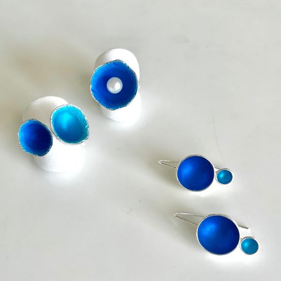 Vier Stücke blau emaillierter Schmuck auf einer weißen Oberfläche. Ein Paar Ohrringe mit einem großen blauen Kreis und einem kleineren blauen Kreis darunter. Ein anderes Stück ist ein Paar Ohrringe mit einer Perle in einem blauen Kreis.