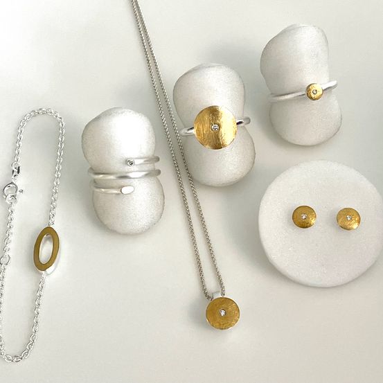 Eine Sammlung von Silber- und Goldschmuck auf einer weißen Oberfläche. Enthält Ringe, eine Halskette und eine Armband. Der Schmuck hat ein modernes und elegantes Design.