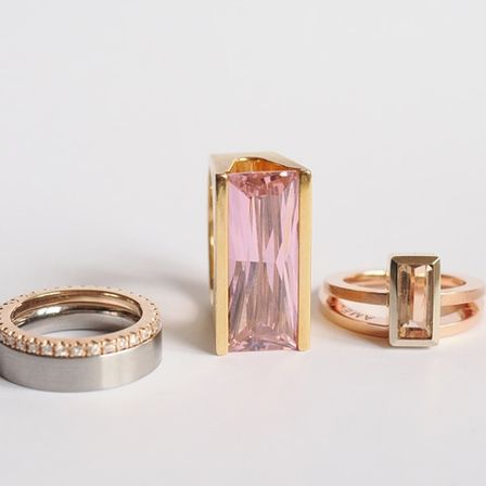 Drei Ringe sind auf einem weißen Hintergrund zu sehen: ein Goldring, ein Roségoldring und ein Silberring. Der mittlere Ring ist größer mit einem rosa Edelstein.