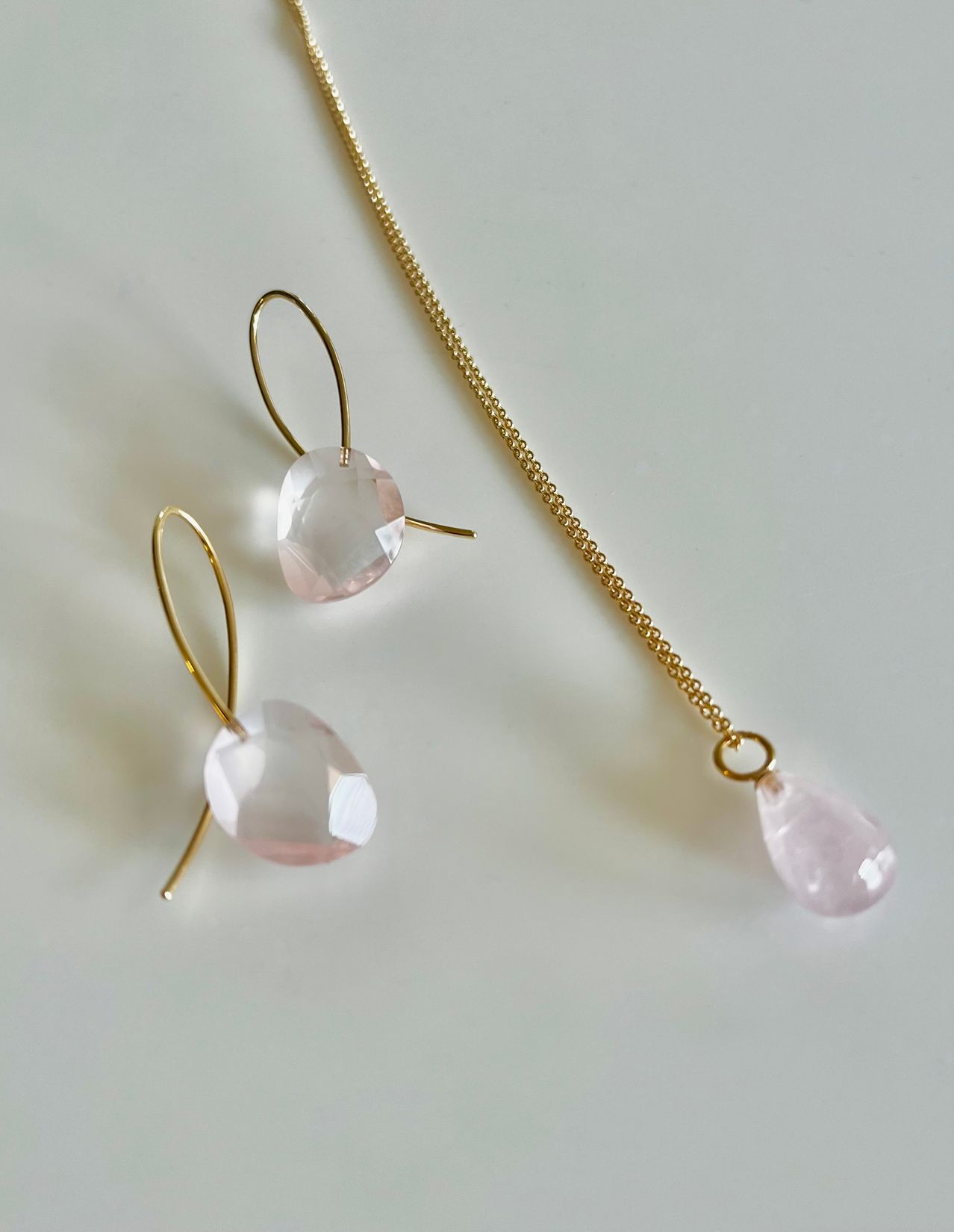 Ein Set aus vergoldeten Ohrringen und einer Halskette mit einem zarten rosa Kristallanhänger auf weißem Hintergrund.