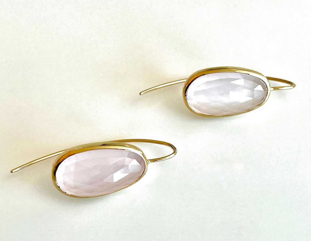 Zwei goldbeschichtete Ohrringe mit ovalen Rosenquarz-Edelsteinen liegen auf einer weißen Oberfläche. Die Steine sind glatt und haben einen zarten rosa Farbton.