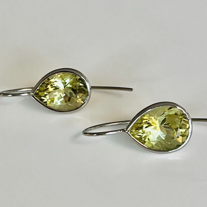 Zwei silberfarbene Ohrringe mit birnenförmigen gelben Edelsteinen werden auf einer weißen Oberfläche präsentiert. Die Edelsteine haben einen hellgelben Farbton und sind gut poliert.