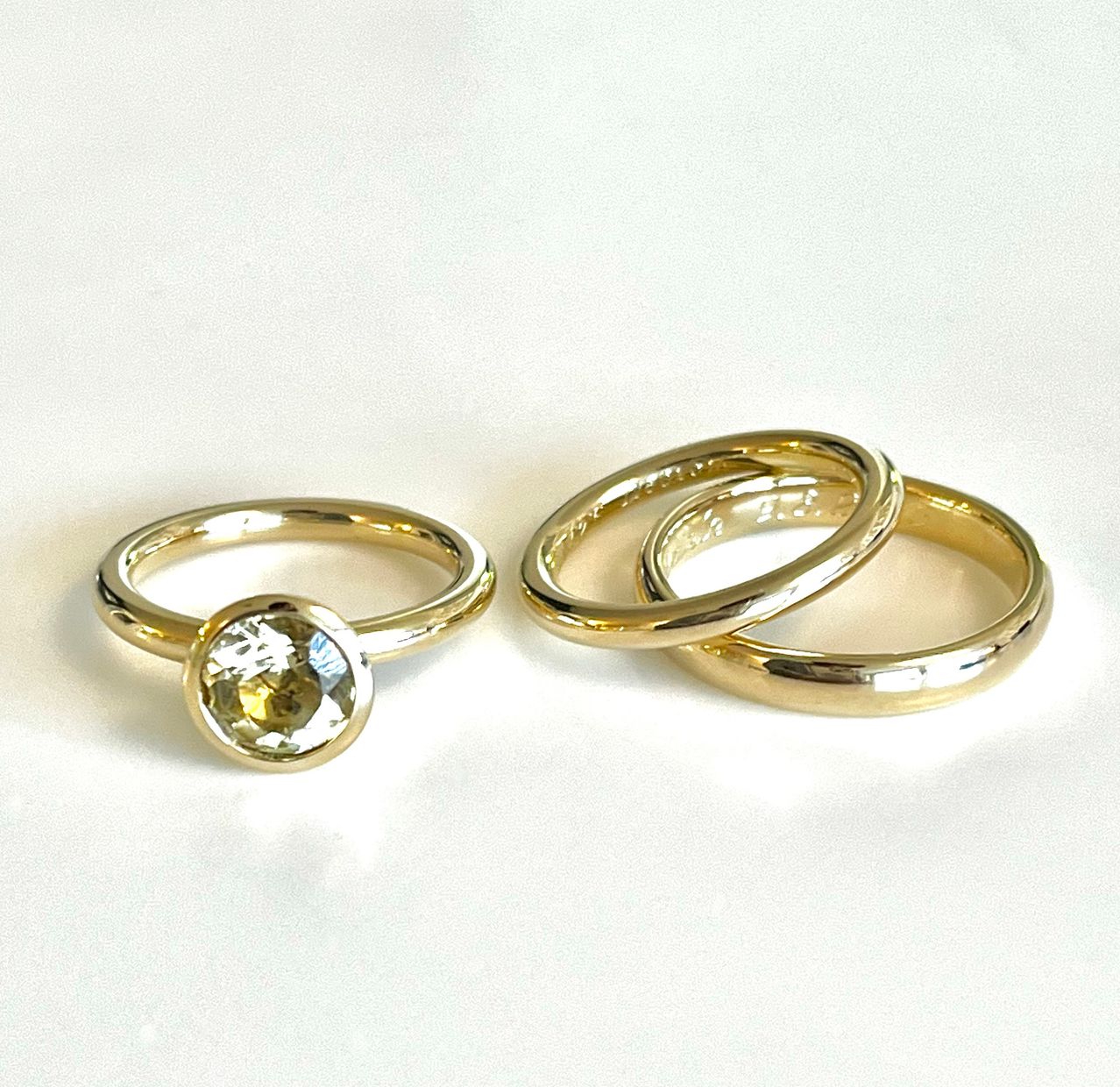 Drei goldene Ringe auf einer weißen Oberfläche, einer mit einem runden gelben Edelstein und zwei schlichten Bändern.