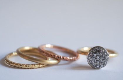 Vier Ringe mit verschiedenen Oberflächen und einem mit Diamanten besetzten Ring auf einer weißen Oberfläche. Der oberste Ring ist aus Gold, der mittlere aus Roségold und der unterste aus Gelbgold.