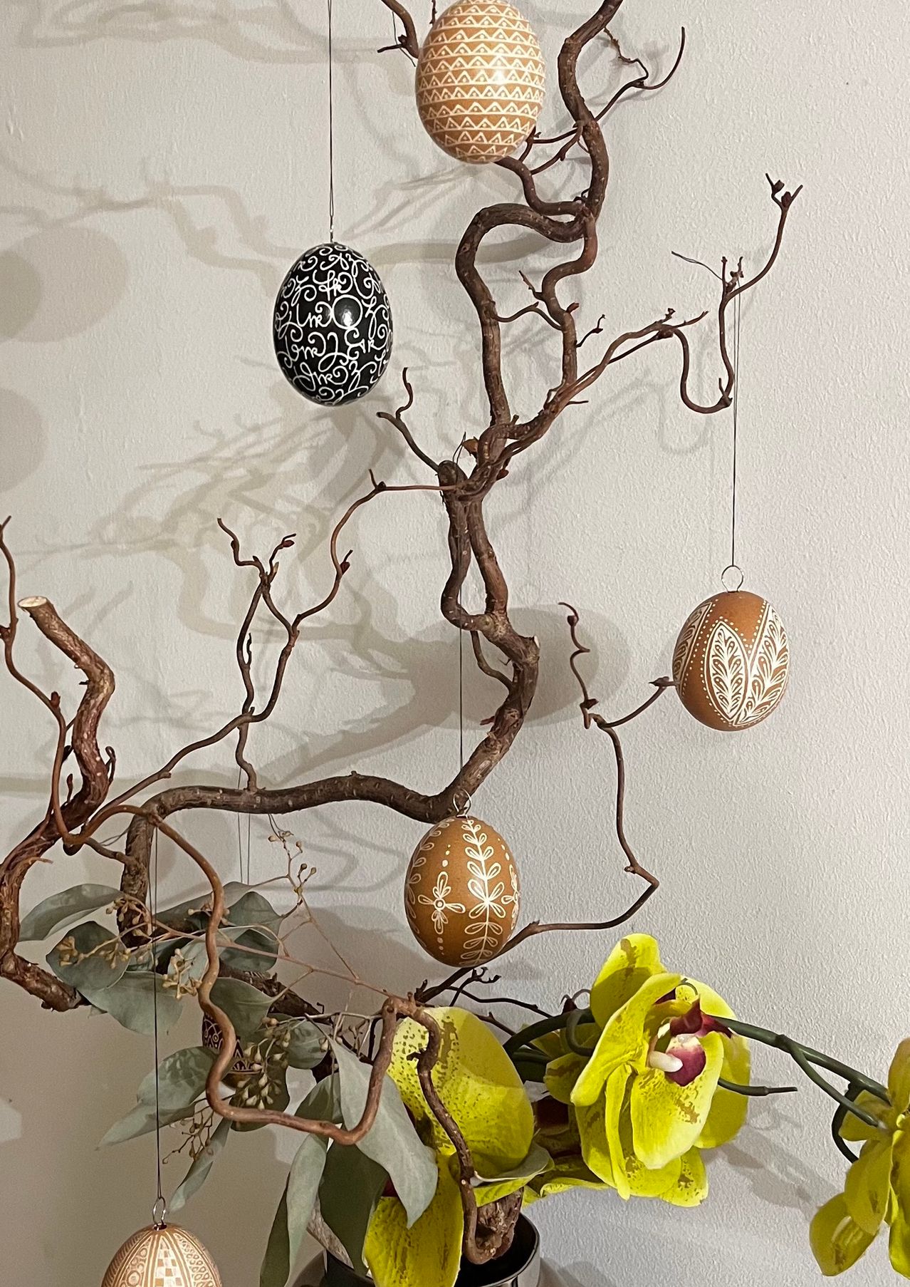 Eine wandmontierte Ausstellung mit einem Baumzweig, an dem drei dekorative Eier hängen, sowie eine gelbe Orchidee. Die Eier haben komplizierte Muster, und der Hintergrund ist eine hellbeige Wand.