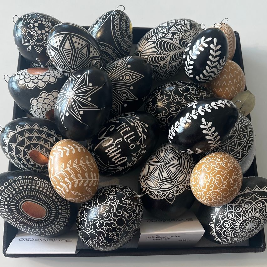 Ein Tablett gefüllt mit kunstvoll verzierten Ostereiern in verschiedenen Designs, einschließlich schwarz mit weißen Mustern und braun mit goldenen Akzenten. Die Eier haben Schnüre zum Aufhängen.
