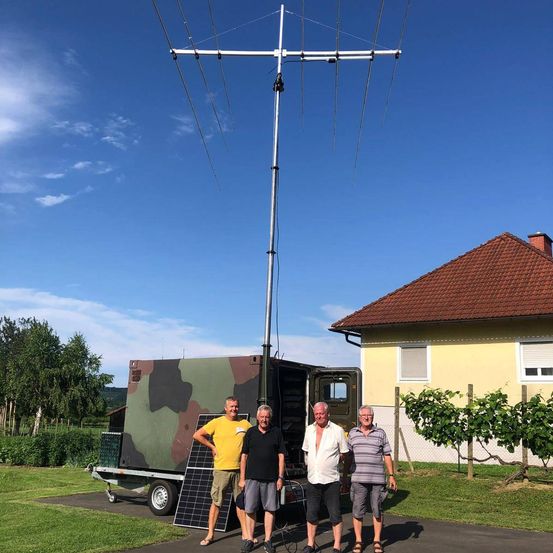 Vier Männer stehen vor einem getarnten Anhänger mit Solarmodulen. Dahinter befindet sich eine große Antenne. Das Haus dahinter hat ein braunes Dach und gelbe Wände.