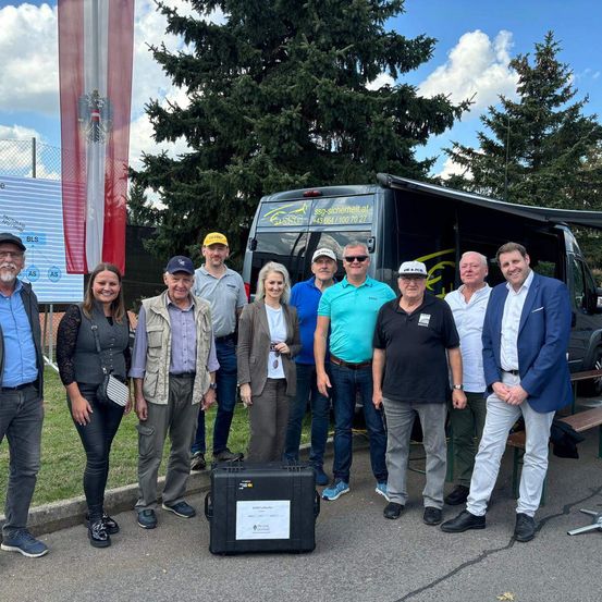Eine Gruppe von Menschen steht vor einem Van und lächelt für ein Foto. Hinter ihnen befinden sich Bäume und eine Fahne.