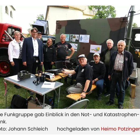 Eine Gruppe von Menschen versammelt sich um ein getarntes Militärfahrzeug, möglicherweise für eine Veranstaltung. Ein Mann sitzt auf einer Bank mit einem Laptop, und ein anderer Mann steht neben einem Tisch mit verschiedenen Geräten.