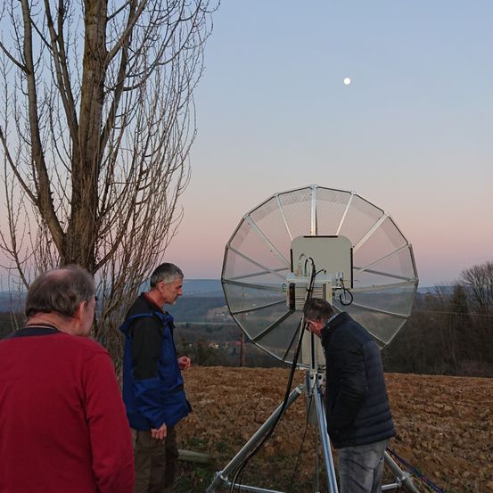 Drei Männer stehen um eine große Satellitenschüssel herum. Die Schüssel steht auf einem Stativ, und ein Mann schaut darauf. Sie sind draußen auf einem Feld bei einem mondhellen Himmel.