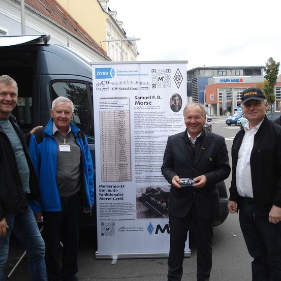 Vier Männer stehen vor einem Banner mit Morse-Code-Details und einem Fahrzeug im Hintergrund. Der Mann in der Mitte hält ein kleines elektronisches Gerät.