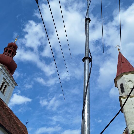 Ein hoher Metallmast steht zwischen zwei Kirchtürmen. Die Türme haben rote Dächer und weiße Wände. Der Himmel ist blau mit weißen Wolken.