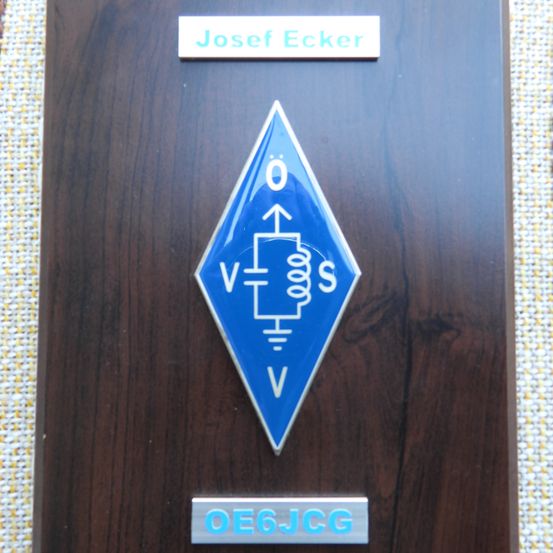 Eine hölzerne Plakette mit einem blauen Rautensymbol, eingraviert mit einem Spannungssymbol und dem Namen 'Josef Ecker' darüber. Unten steht das Rufzeichen 'OE6JCG'.