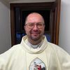 Ein Priester steht lächelnd in einem Raum und trägt ein weißes Gewand mit einem Kreuz und einem Bild auf der Vorderseite. Hinter ihm befindet sich eine Wand mit einer Uhr.