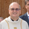 Ein lächelnder Mann in einem weißen geistlichen Gewand mit einem goldenen Kreuz steht neben einem Mann in einem Anzug. Der Hintergrund zeigt eine Holzwand.