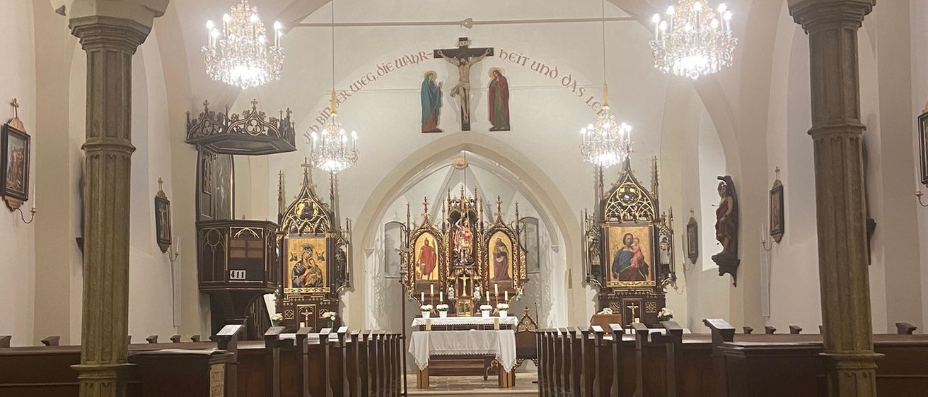 Der Innenraum einer Kathedrale mit hölzernen Kirchenbänken, einem Altar und einem Kruzifix, geschmückt mit Kronleuchtern und religiösen Kunstwerken.