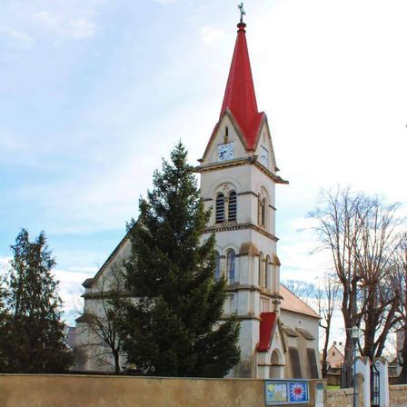 Eine weiße Kirche mit einem roten Kirchturm steht hoch gegen einen blauen Himmel, umgeben von grünen Bäumen. Eine Uhr befindet sich auf dem Kirchturm.