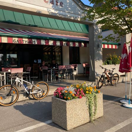 Ein Outdoor-Cafe mit einem rot-weiß gestreiften Vordach. Zwei Fahrräder sind davor geparkt, und es gibt ein Blumenbeet mit bunten Blumen. Zwei Personen sitzen an einem Tisch draußen.