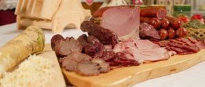 Ein Holzschneidebrett mit verschiedenen Fleischsorten wie Schinken, Würsten und Salami. Andere Gegenstände auf dem Tisch sind ein Holztablett und einige Gemüse.