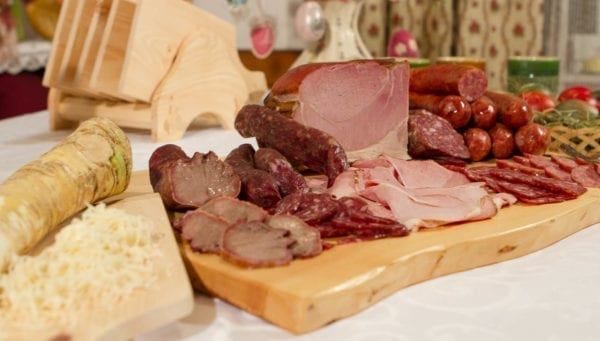 Ein Holzschneidebrett mit verschiedenen Fleischsorten wie Schinken, Würsten und Salami. Andere Gegenstände auf dem Tisch sind ein Holztablett und einige Gemüse.