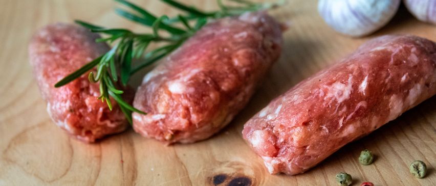 Auf einem Holzschneidebrett befinden sich drei rohe Wurststücke, verziert mit Rosmarinzweigen und begleitet von einer Knoblauchzehe und kleinen Pfefferkörnern.