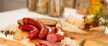 Ein Holzschneidebrett mit mehreren Würsten, geschnittenem Schinken und geriebenem Käse. Daneben liegen ein Messer und eine Gabel. Dahinter sind verschiedene Gläser mit Lebensmitteln zu sehen.