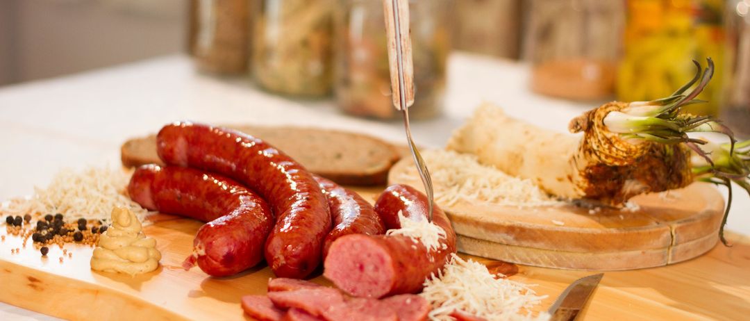 Ein Holzschneidebrett mit mehreren Würsten, geschnittenem Schinken und geriebenem Käse. Daneben liegen ein Messer und eine Gabel. Dahinter sind verschiedene Gläser mit Lebensmitteln zu sehen.