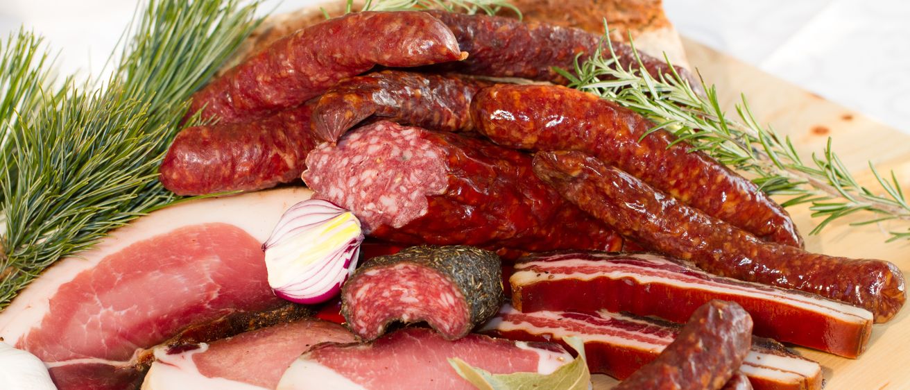 Eine Nahaufnahme verschiedener Wurstwaren, einschließlich Würste, Speck und Salami, auf einem Holzbrett mit Kräutern und Gewürzen.