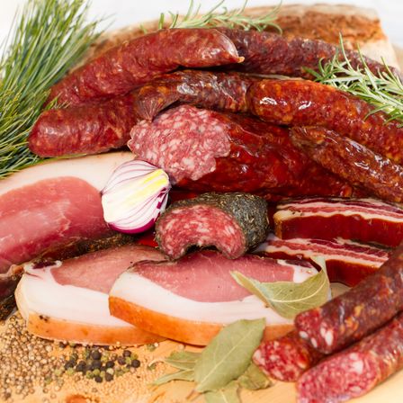 Eine Auswahl an Wurstwaren wie Würste, Salami und Speck, garniert mit Rosmarin und Zwiebeln, auf einem Holzbrett angeordnet.