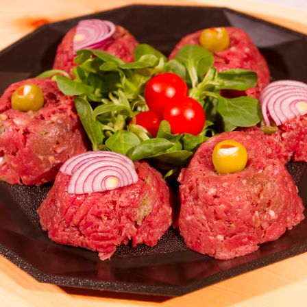 Ein schwarzer Teller zeigt sechs Rinderpatties mit grünen Oliven und roten Zwiebeln, dazu ein Salat aus Tomaten und grünem Blattsalat.