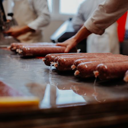 Arbeiter in einer Küche handhaben Würste, einige in Plastik verpackt. Die Würste liegen auf einem Edelstahltisch.