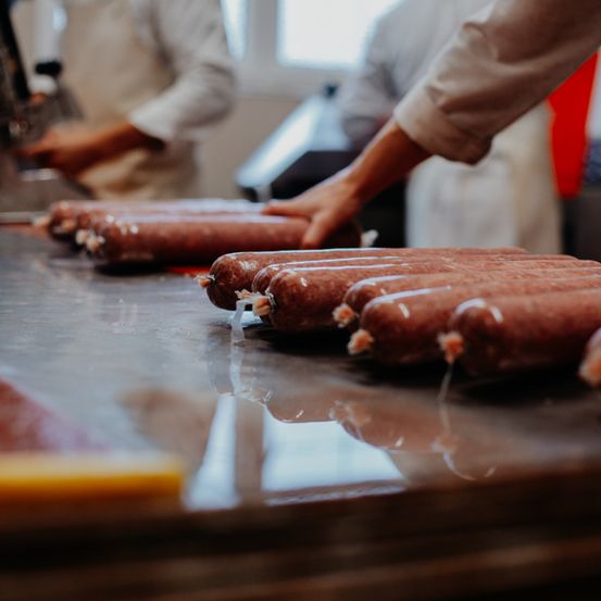 Arbeiter in einer Küche handhaben Würste, einige in Plastik verpackt. Die Würste liegen auf einem Edelstahltisch.