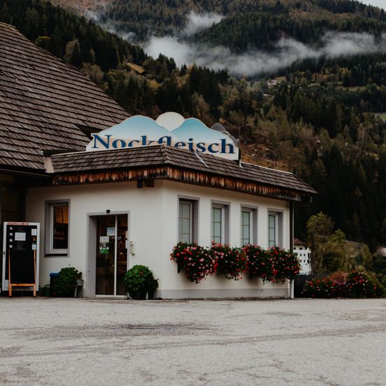 Ein Gebäude mit einem braunen Dach und einem Schild mit der Aufschrift 'Nock-Aleisch'. Es hat Glasfenster und eine Tür. Das Gebäude ist von Blumen und Pflanzen umgeben. Im Hintergrund befindet sich ein Berg mit Nebel.