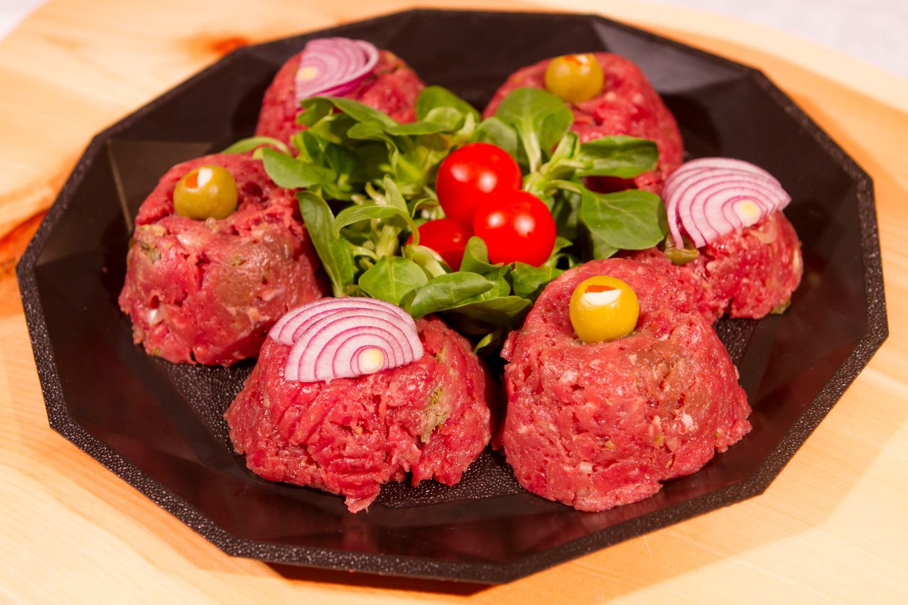 Ein Teller mit sechs Rinderpatties, jedes belegt mit einer grünen Olive und einer Scheibe roter Zwiebel. Grüner Salat mit Tomaten ist ebenfalls auf dem Teller.