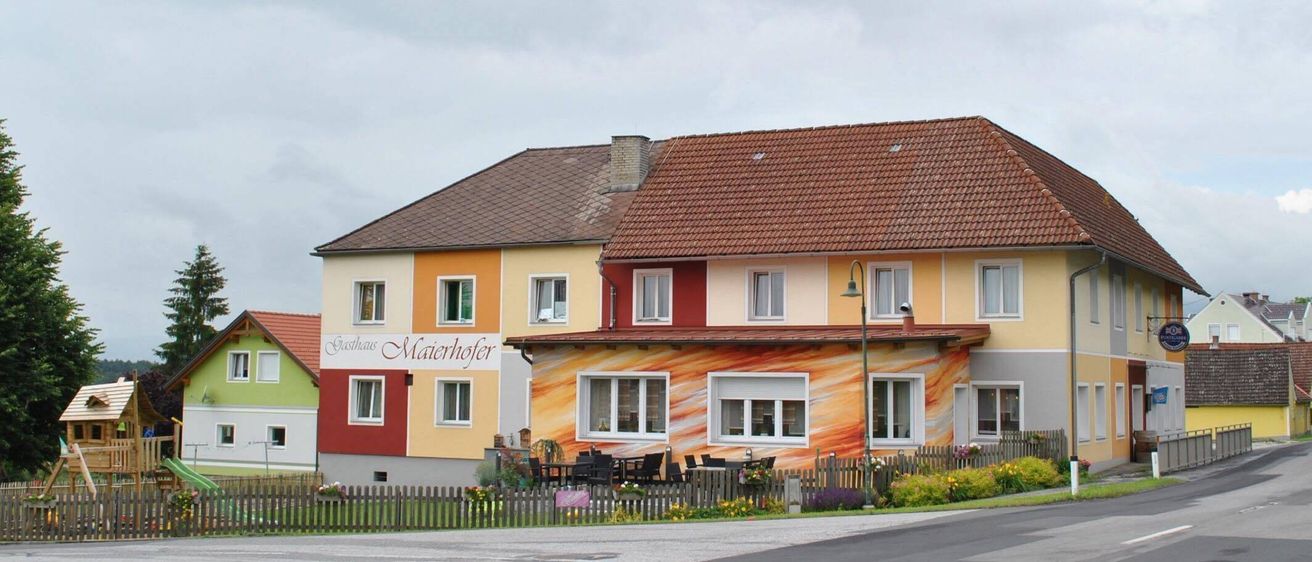 Ein Haus mit einem Ziegeldach, in Gelb, Orange und Rot gestrichen, mit dem Namen Maierhofer. Vor dem Haus stehen Tische und Stühle.